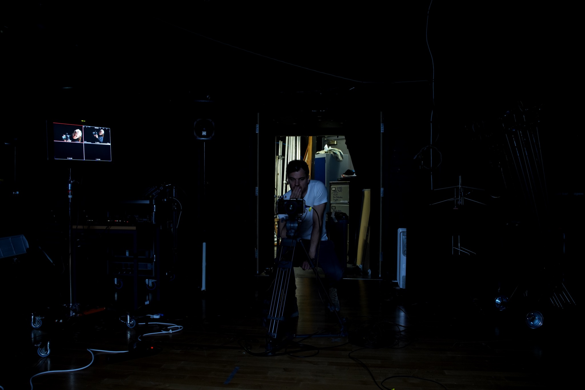 The director crouched behind a cinema camera in the dark studio, a monitor glowing to one side.