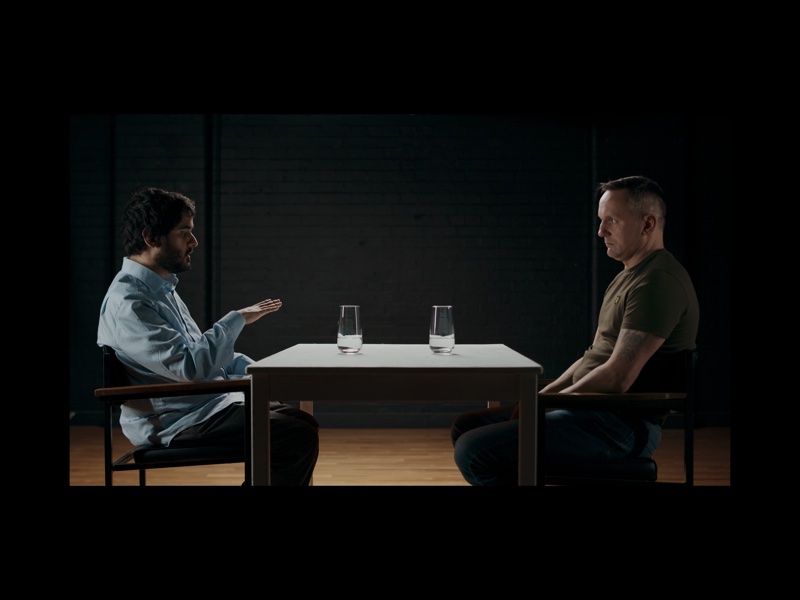 Two men face each other across a small table, in front of a black brick wall.